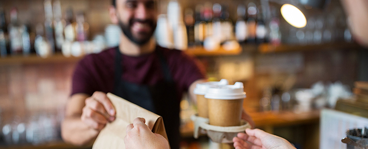 Blurred image of a barista handing two cups of coffee and a brown paper bag to a customer who is out of shot.